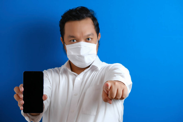 Maske takan şişman Asyalı ofis çalışanı akıllı telefonunu ciddi bir bakışla işaret ediyor. işaret etti. mavi arkaplan - Fotoğraf, Görsel
