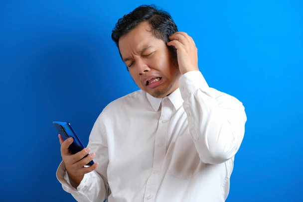Asiatischer Geschäftsmann beim SMS-Chatten auf seinem Handy, schlechte Nachrichten, traurige weinende Miene. blauer Hintergrund - Foto, Bild