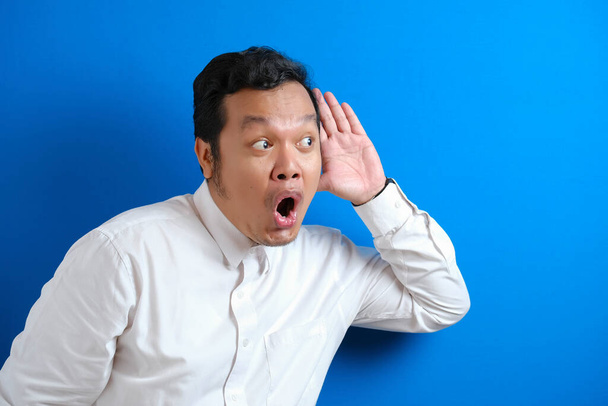 Young asian businessman wearing white shirt listen whispers with funny face. Curious. Intently listen in on juicy gossip. Conversation news, privacy violation, against blue wall - Photo, Image