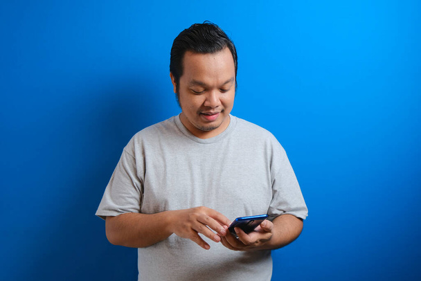 φωτογραφία ενός χοντρού Ασιάτη με γκρι μπλουζάκι που δείχνει χαρούμενος ενώ έχει πρόσβαση σε καλά νέα στο smartphone του. οι άνδρες δείχνουν αυτοπεποίθηση χειρονομίες. απομονωμένο σε μπλε φόντο - Φωτογραφία, εικόνα