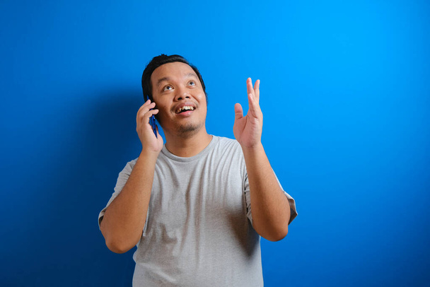Telefon alırken gülümseyen gri tişörtlü şişman bir Asyalı adamın fotoğrafı. Mavi arkaplanda izole edilmiş - Fotoğraf, Görsel