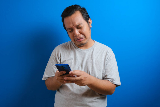Толстый азиат в серой футболке выглядит грустным, читая онлайн новости со своего сотового. мужчина показывает разочарованный жест, вытирая слезы с лица
 - Фото, изображение