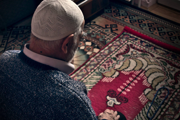 Velmi starý vousatý turecký muslimský muž v osmdesátých letech nosí modlitební čepici a modlí se u sebe doma na modlitebním koberci v ramadánu. - Fotografie, Obrázek