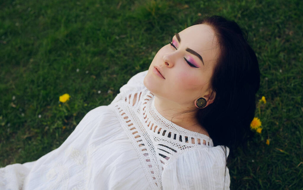 Ta dívka leží na trávě v parku. Krásný růžový make-up na dívce. Bruneta nevěsta v krásných boho šaty, s pozadím na poli s květinami. Jemný portrét v bílých šatech. Šik černé vlasy s dlouhými vlasy. - Fotografie, Obrázek