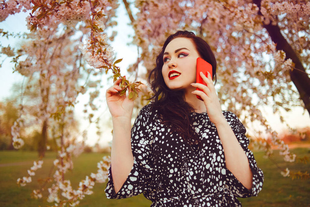 Egy lány a parkban egy fa hátterében virágokkal, virágokkal. A lány telefonon beszél, és a virágok közelében sétál a természetben. Barna hajú, piros ajkakkal és egy pálcával a telefonhoz.. - Fotó, kép