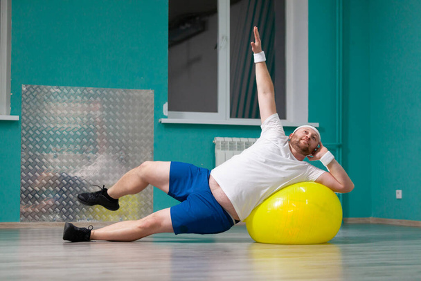 Χαμογελώντας λίπος άνθρωπος κάνει ασκήσεις χρησιμοποιώντας μπάλα γυμναστικής. Υπέρβαρος άνθρωπος είναι ευχαριστημένος με το αποτέλεσμα της εκπαίδευσής του σε ομαδικά μαθήματα φυσικής κατάστασης - Φωτογραφία, εικόνα