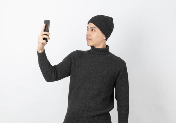 Akıllı telefon oynarken gri kazak ve bere giyen yakışıklı Asyalı genç adam beyaz arka planda selfie çekiyor.. - Fotoğraf, Görsel