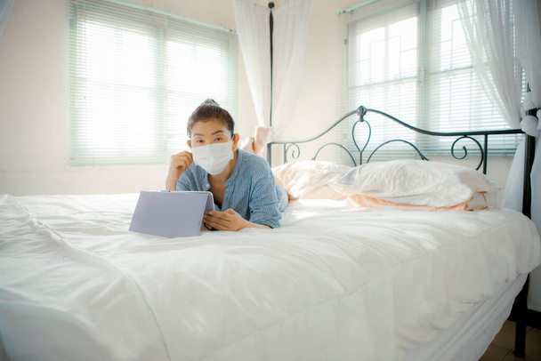 Elinde koruma maskesi olan bir kadın evdeki oturma odasında kanepeye uzanmış internet cihazıyla sohbet ediyor. - Fotoğraf, Görsel