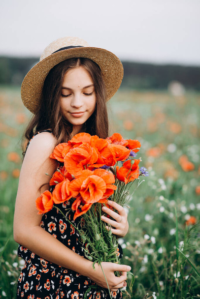 Beautiful young free girl in a hat in a summer field of red poppies with a bouquet. Soft selective focus. - Photo, Image