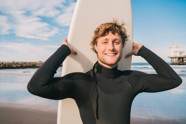 Close-up of young surfer standing at the beach with his surfboard and wearing black surfing suit. Sport and water sport concept. - Foto, Bild