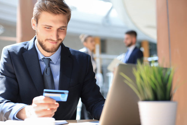 Smiling man sitting in office and pays by credit card with his laptop - Foto, Imagen