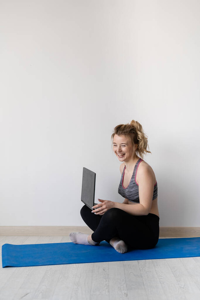 Glückliche junge Frau sitzt mit überkreuzten Beinen auf dem Boden und benutzt Laptop auf grauem Hintergrund - Foto, Bild