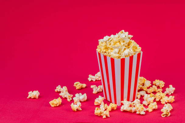 Papierbecher mit Popcorn auf farbigem Hintergrund. Gestreifte Schachtel mit Popcorn auf rotem Hintergrund. Popcorn im rot-weiß gestreiften Pappeimer . - Foto, Bild