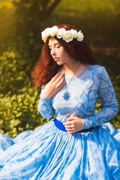 Portrait of an attractive woman with a rim of white flowers and in a blue magnificent dress sitting on the grass in the park against the background of lilac bushes.  - Photo, image