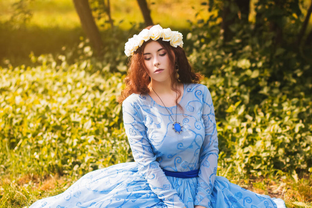 Πορτρέτο μιας ελκυστικής γυναίκας με ένα χείλος από λευκά λουλούδια και σε ένα μπλε υπέροχο φόρεμα κάθεται στο γρασίδι στο πάρκο με φόντο λιλά θάμνους.  - Φωτογραφία, εικόνα