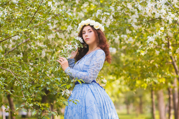 Een mooie vrouw in een blauwe gezwollen jurk met een rand van witte bloemen op haar hoofd loopt door het park tussen bloeiende appelbomen. - Foto, afbeelding