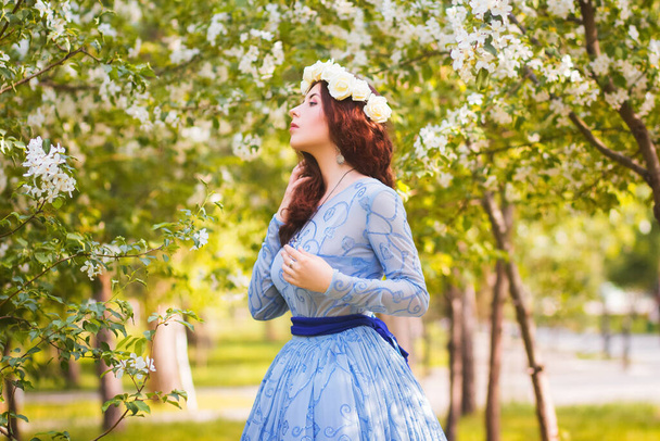 A beautiful woman in a blue puffy dress with a rim of white flowers on her head walks through the park between blooming apple trees. - Фото, изображение