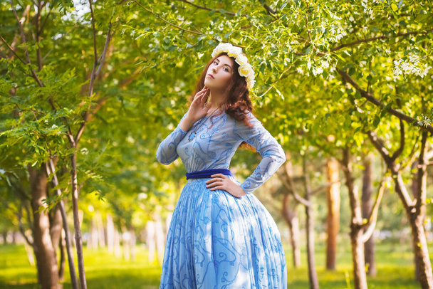 A beautiful woman in a blue puffy dress with a rim of white flowers on her head walks through the park between blooming apple trees. - Фото, изображение