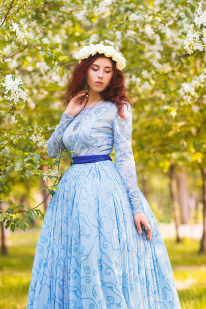 A beautiful woman in a blue puffy dress with a rim of white flowers on her head walks through the park between blooming apple trees. - Фото, зображення