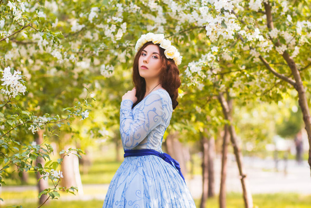 Een mooie vrouw in een blauwe gezwollen jurk met een rand van witte bloemen op haar hoofd loopt door het park tussen bloeiende appelbomen. - Foto, afbeelding