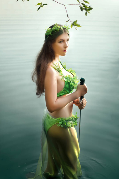 An attractive woman in a swimsuit of green plants with a wreath on her head stands knee-deep in troubled water holding a sword in her hands. Concept art of a mistress of the lake from legends. - Photo, Image
