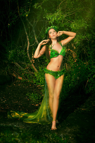 Attractive woman in a swimsuit of green plants with a wreath on her head posing while standing on a wet shore against a background of thickets. Concept art of a mistress of the lake from legends. - Photo, Image