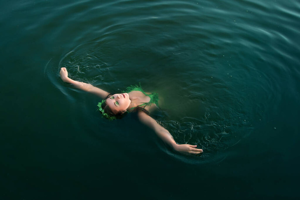 Una bella ragazza in costume da bagno di piante verdi con una ghirlanda sulla testa galleggia sulla schiena in acque agitate. Concetto arte di un'amante del lago da leggende
. - Foto, immagini
