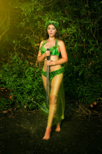 Attractive woman in a swimsuit of green plants with a wreath on her head holds a sword in her hands. Concept art of a mistress of the lake from legends.  - Фото, зображення