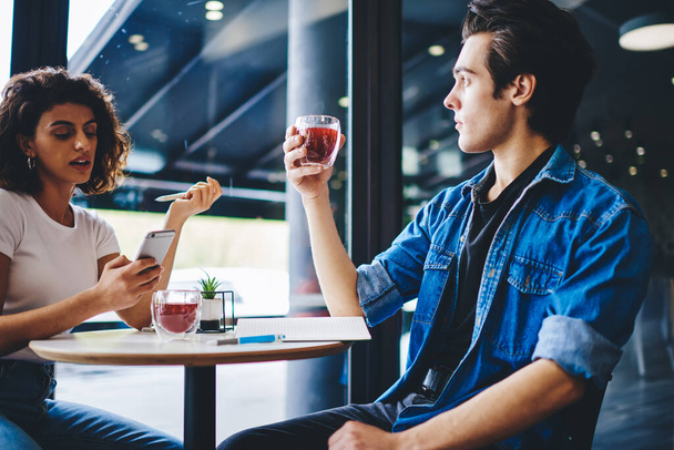 Young woman chatting in social network on smartphone while spending leisure time together with boyfriend in coffee shop interior.Stylish dressed couple in love drinking tasty juice in cafe - Photo, Image