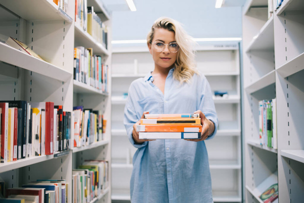 Serious young woman in casual wear standing near bookshelves holding literature for education in college, caucasian female librarian in optical spectacles offering books for reading on leisure - Photo, image