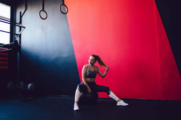 Contemplative female athlete in trendy tracksuit sitting at swiss ball in modern studio and waiting start of cardio training, thoughtful woman in active wear holding hair and looking away in gym - Foto, imagen