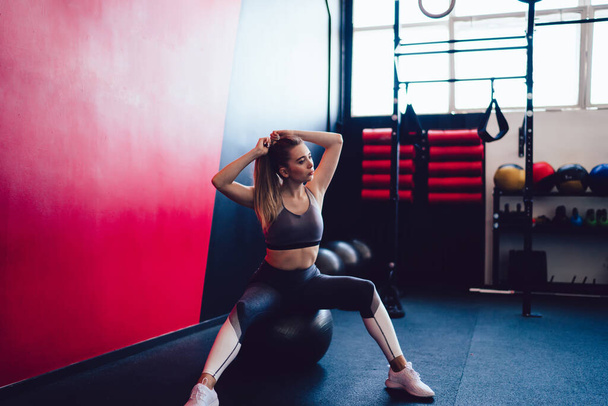 Caucasian fitness woman dressed in stylish tracksuit resting on pilates swiss ball in modern gym studio, attractive physical female athlete spending time for healthy lifestyle day in sport hall - Zdjęcie, obraz