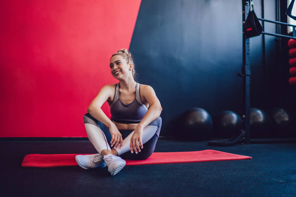 Pozytywny hipster dziewczyna ubrana w stylowe dresy uśmiechnięta podczas odpoczynku na czerwonym dywanie do treningu pilates, szczęśliwy sportowiec kobiet korzystających z czasu na crossfit w hali sportowej uczucie beztroski - Zdjęcie, obraz