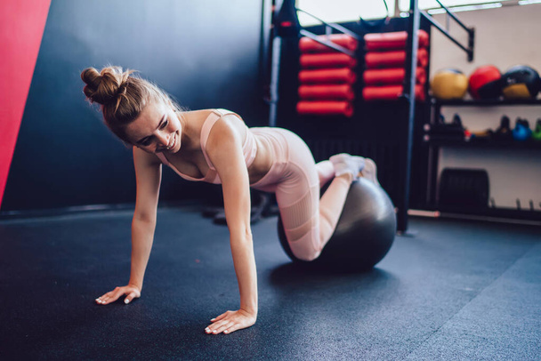 Cheerful Caucasian hipster girl in trendy tracksuit looking away and smiling during cardio exercising in gym, positive female athlete feeling good during training with swiss ball in pilates studio - Foto, immagini