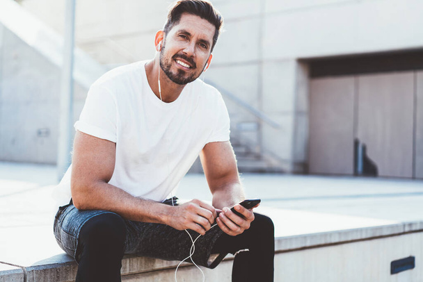 Concept of healthy lifestyle and time for cardio training outdoors, portrait of handsome sportsman in headphones looking at camera while taking rest on urban setting after morning running in city - Fotoğraf, Görsel