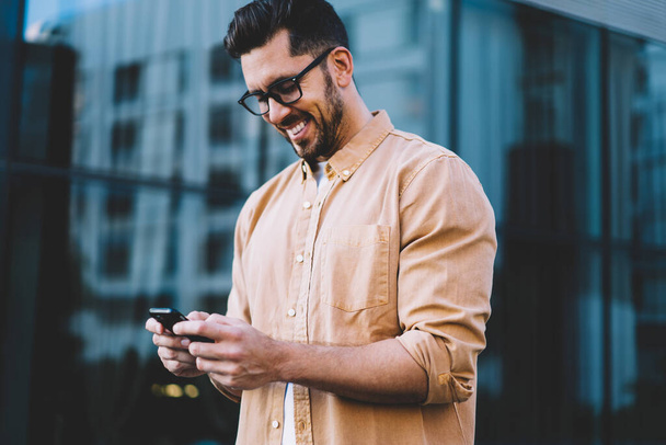 Successful bearded man in eyeglasses for provide eyes protection smiling while reading positive sms from friend connected to 4g,happy male browsing website via smartphone gadget while standing outdoor - 写真・画像