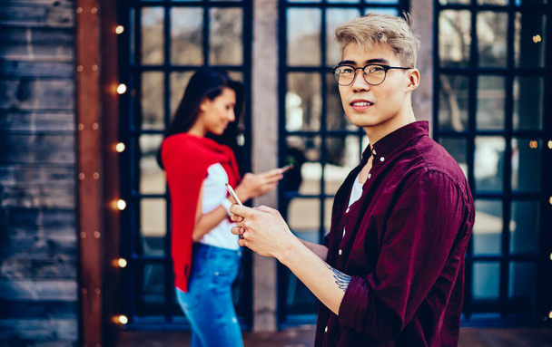 Pondering chinese hipster guy in eyeglasses looking away while messaging on smartphone and using 4G internet for communication with friend which standing on blurred background with cellular in hand - 写真・画像