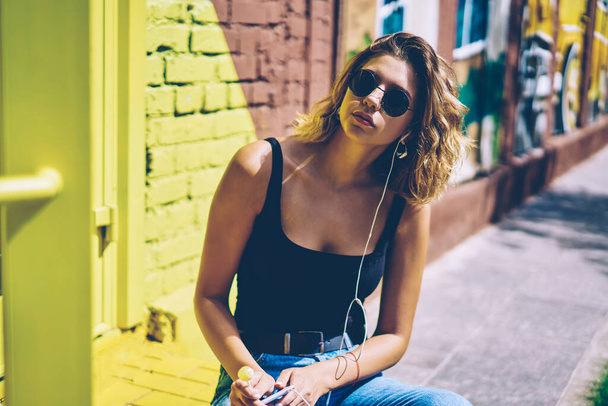 Portrait of confident female meloman in sunglasses looking at camera while listening music from popular radio station via electronic equipment, young hipster girl in headphones enjoying summer weather - 写真・画像