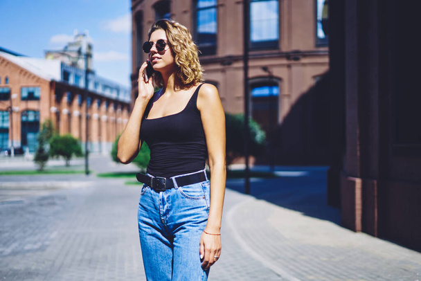 Pretty hipster girl using roaming connection for communicate with friends via cellular phone during walking around summer city, millennial woman calling to operator for checking account balance - Foto, Bild