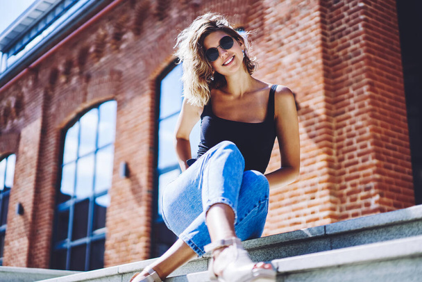 Portrait of cheerful female teenager looking at camera while sitting on stairs at college campus, successful hipster girl in sunglasses enjoying free time for posing on urban city during summer day - 写真・画像