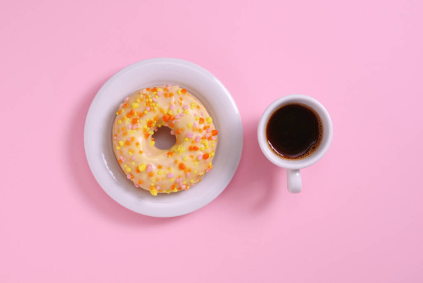 Donut com gelo e polvilhas e xícara de café preto sobre fundo rosa, vista superior
 - Foto, Imagem