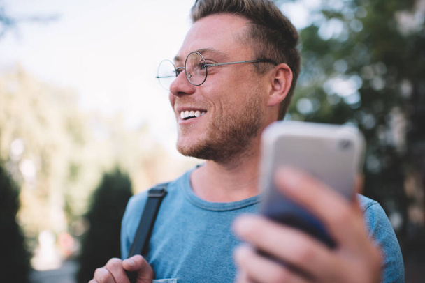 Close side view of podekscytowany wesoły hipster facet w okularach uśmiecha się podczas odwracania wzroku i czeka na telefon komórkowy z obsługi klienta, pozytywny człowiek korzystających spędzając wolny czas trzymając smartfona - Zdjęcie, obraz