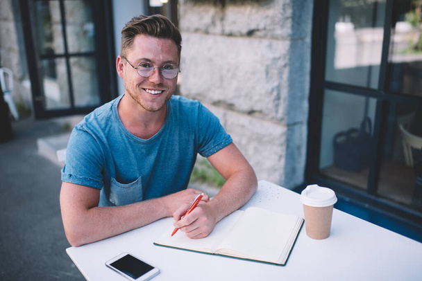 Portrait of happy male student sitting at cafeteria table with textbook for studying and doing college test, cheerful Caucasian man in spectacles for provide eyes protection smiling at camera - 写真・画像
