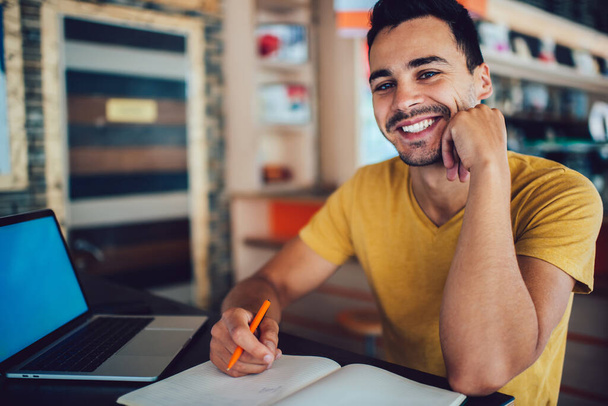 Πορτρέτο του χαρούμενος hipster τύπος κοιτάζοντας κάμερα και χαμογελώντας, ενώ κάθεται στην επιφάνεια εργασίας με φορητό υπολογιστή για την ηλεκτρονική μάθηση και το βιβλίο για την εκπαίδευση της γνώσης, χαρούμενα αρσενικό φοιτητής αισθάνεται καλά - Φωτογραφία, εικόνα