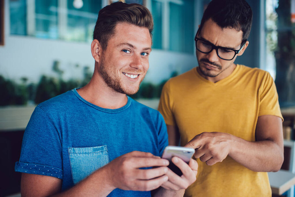 Half length portrait of cheerful Caucasian male blogger smiling at camera while spending spare time with best friend, millennial man checking email box via application on smartphone gadget - Valokuva, kuva