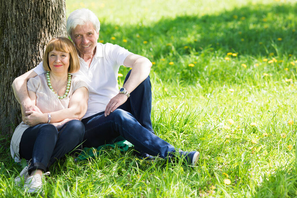 Ελκυστικό παντρεμένο ηλικιωμένο ζευγάρι απολαμβάνοντας μαζί κάθεται κάτω από το δέντρο στο λιβάδι άνοιξη - Φωτογραφία, εικόνα