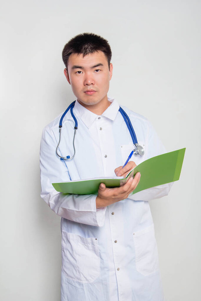A young Asian doctor in a white coat, with a stethoscope on his neck, holds a green folder in his hands and takes notes. One on a white background. - Foto, imagen