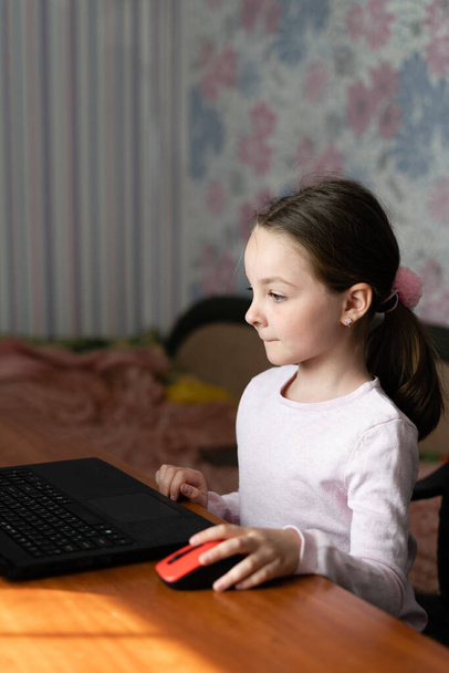 little ethnic girl sitting at home at a table working on her homework on a laptop computer reading the screen with a thoughtful expression - Photo, Image