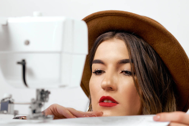 Cute girl in hat uses sewing machine sews paper - Zdjęcie, obraz