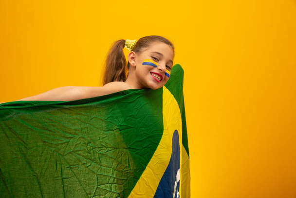 Υποστηρικτής ποδοσφαίρου, ομάδα Βραζιλίας. Παγκόσμιο Κύπελλο. Όμορφο κοριτσάκι ζητωκραυγάζει για την ομάδα της σε κίτρινο φόντο - Φωτογραφία, εικόνα